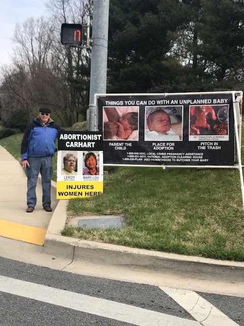 Pray for Bethesda has 	posted signs alongside sidewalks on Old Georgetown Road and the group plans keep protesting outside all winter. Here, group member Ken Garrison stands next to signs against Wildwood Medical Center abortionist Leroy Carhart. 

