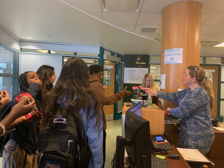 The attendance secretary handing a student late to fifth-period a pink unexcused tardy pass from the main office. While the policy has good intentions, it does make students miss more time from class especially those who arent repeat tardies.